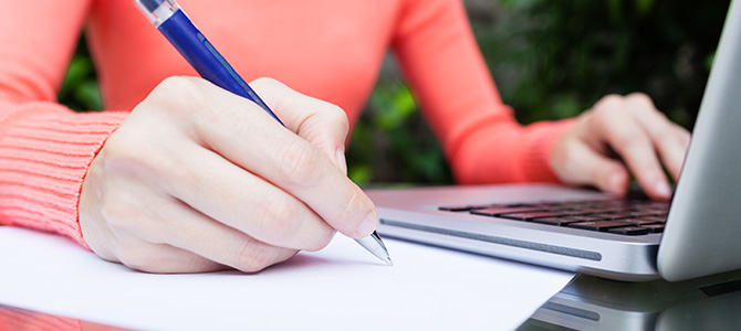 Mulher sentada num jardim, escreve uma carta de demissão enquanto vê informação no seu portátil