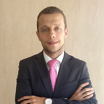 Homem com terno escuro e gravata rosa, braços cruzados.