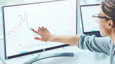 Mulher analisando gráficos financeiros em dois monitores.