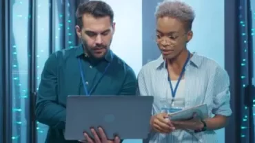 Dois profissionais analisando informações em um laptop em uma sala de servidores.