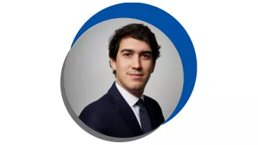 Homem em terno escuro com gravata azul, fundo cinza.