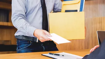 Um colaborador vestido com uma camisa e gravata, entrega uma carta de demissão ao chefe enquanto segura numa caixa com documentos