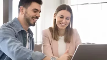 Duas pessoas sentadas à mesa, em frente a um computador portátil, a sorrir durante um momento de integração profissional.
