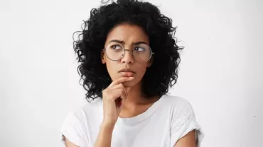 Mulher de óculos pensando, mão no queixo.