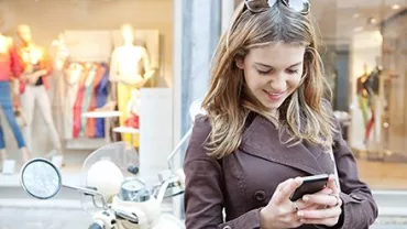 Mulher usando celular na rua, ao lado de uma scooter, frente a uma loja.