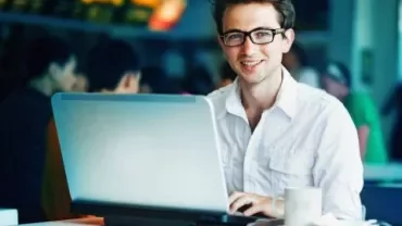 Homem sorrindo enquanto usa laptop em uma cafeteria.