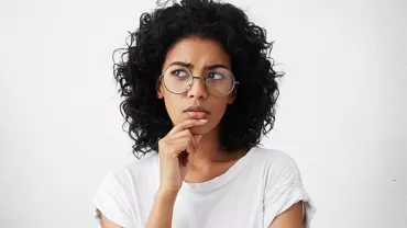 Mulher com cabelo cacheado e óculos, expressão pensativa.