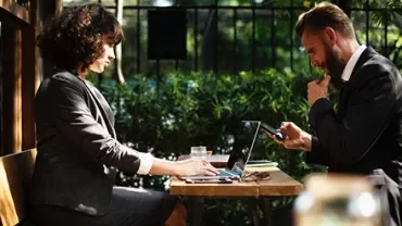 Duas pessoas em uma reunião ao ar livre com laptops e celulares.