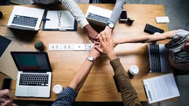 Pessoas reunidas com laptops na mesa, sobrepondo as mãos.