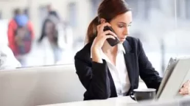 Mulher de terno falando ao celular e usando tablet em um café.