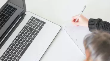Pessoa escrevendo em caderno ao lado de um laptop.