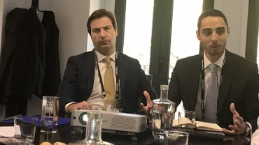 Dois homens de terno em reunião com projetor sobre a mesa.