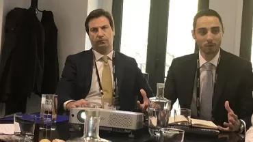 Dois homens de terno em reunião com projetor sobre a mesa.