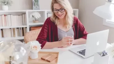 Mulher sorrindo em frente a um laptop em casa com flores na mesa.