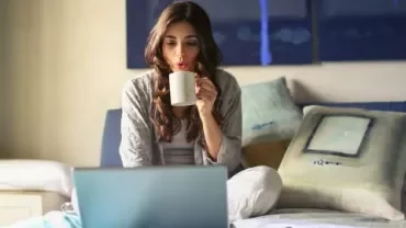 Mulher sentada na cama, bebendo café e usando laptop.
