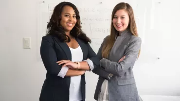 Duas mulheres em roupa formal, sorrindo na frente de um quadro branco.