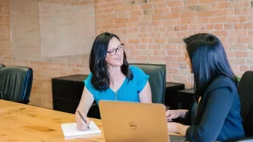 Duas mulheres conversando e sorrindo em uma mesa de reunião.