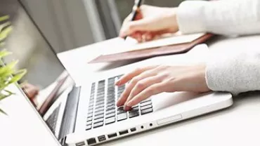 Pessoa digitando em um laptop prateado e escrevendo em um caderno.