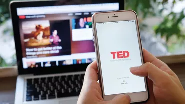 Pessoa segurando um celular com o aplicativo TED aberto, e um laptop ao fundo.