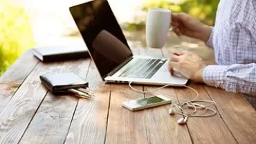 Pessoa com laptop e caneca em mesa de madeira ao ar livre.