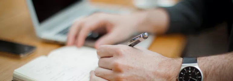 Pessoa escrevendo em um caderno com um laptop ao lado.
