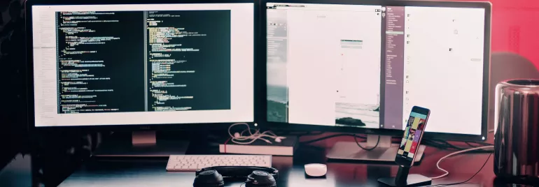 Mesa com dois monitores mostrando código, teclado e mouse.