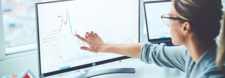 Mulher analisando gráficos financeiros em dois monitores.