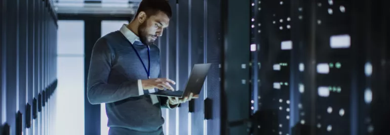 Homem usando laptop em corredor de servidores em data center.