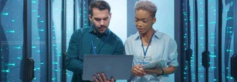Dois profissionais analisando informações em um laptop em uma sala de servidores.