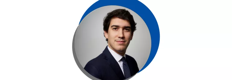 Homem em terno escuro com gravata azul, fundo cinza.