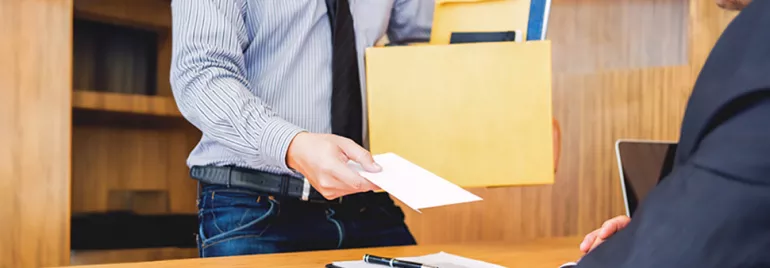 Um colaborador vestido com uma camisa e gravata, entrega uma carta de demissão ao chefe enquanto segura numa caixa com documentos