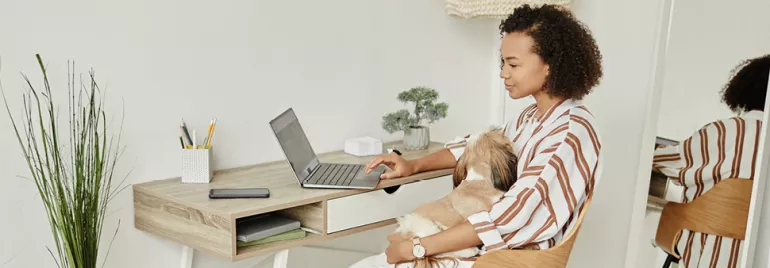 Mulher trabalha no notebook com cachorro no colo em um escritório em casa.