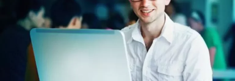 Homem sorrindo enquanto usa laptop em uma cafeteria.