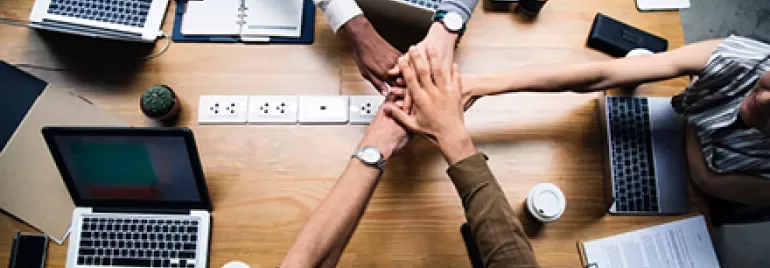 Pessoas reunidas com laptops na mesa, sobrepondo as mãos.