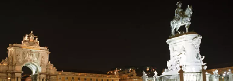 Praça iluminada à noite com estátua e arco ao fundo.