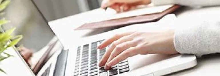 Pessoa digitando em um laptop prateado e escrevendo em um caderno.