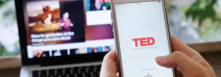 Pessoa segurando um celular com o aplicativo TED aberto, e um laptop ao fundo.
