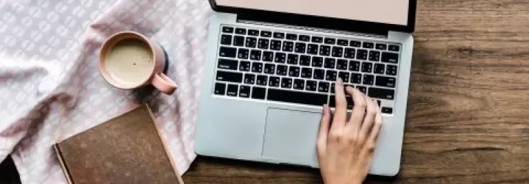 Pessoa usando laptop em mesa de madeira com xícara e caderno.