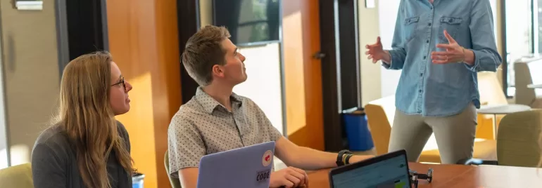 Reunião em escritório com três pessoas conversando, laptops sobre a mesa.