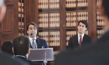 Homem de terno falando em púlpito em biblioteca com prateleiras de livros.