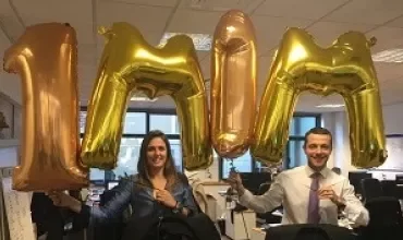 Dois colegas seguram balões dourados formando a palavra '1M' em um escritório.