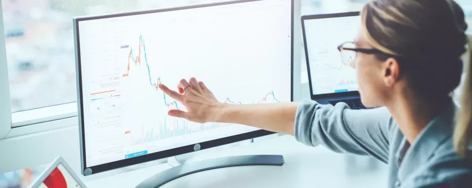 Mulher analisando gráficos financeiros em dois monitores.
