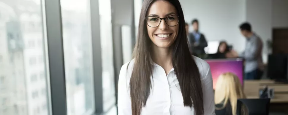 Mulher sorrindo em um escritório moderno com colegas ao fundo.