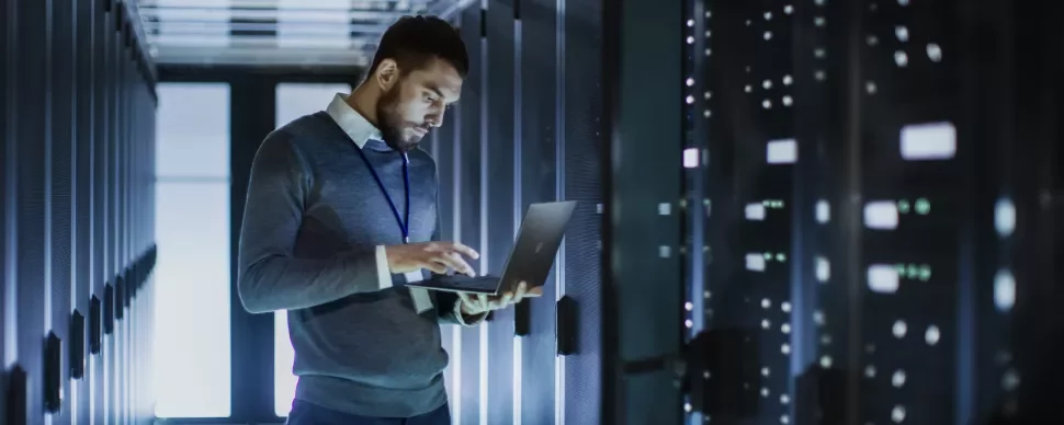 Homem usando laptop em corredor de servidores em data center.