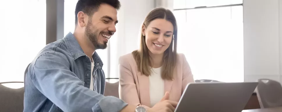 Duas pessoas sentadas à mesa, em frente a um computador portátil, a sorrir durante um momento de integração profissional.