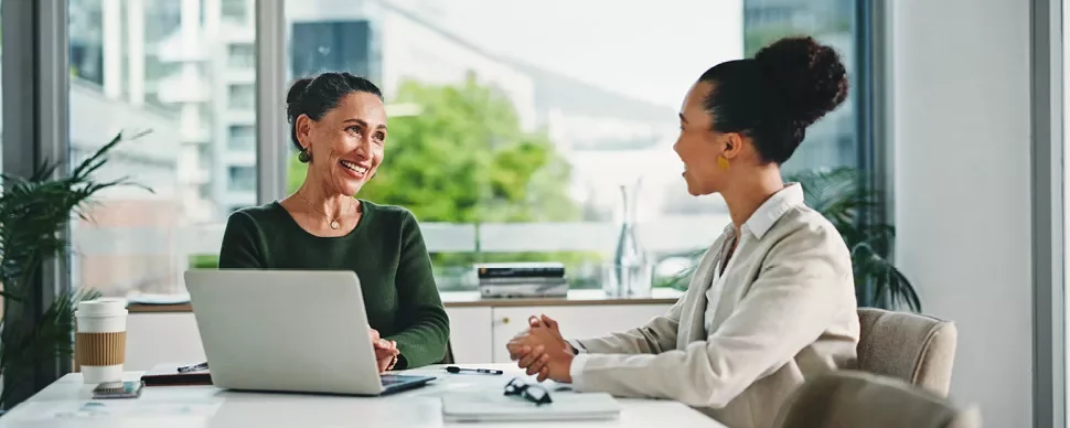 Duas mulheres a conversar, sentadas numa mesa, com um portátil aberto entre ambas. 
