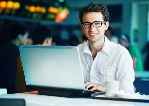 Homem sorrindo enquanto usa laptop em uma cafeteria.