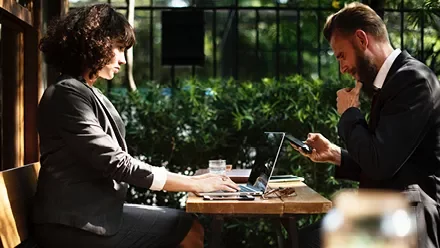 Duas pessoas em uma reunião ao ar livre com laptops e celulares.