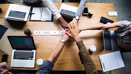 Pessoas reunidas com laptops na mesa, sobrepondo as mãos.