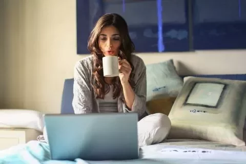 Mulher sentada na cama, bebendo café e usando laptop.